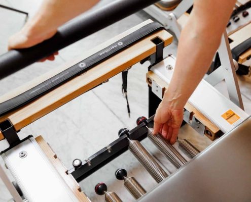 Pilates-Reformer Person trainiert an einem Pilates-Reformer und greift mit einer Hand an die Federn des Geräts.