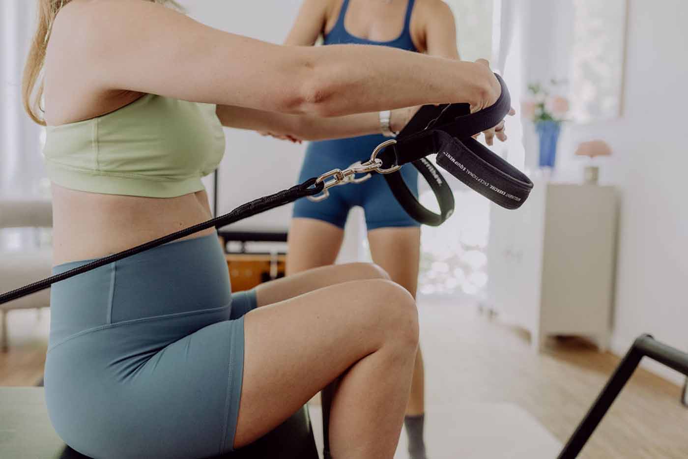 Pilates-Übung am Reformer Nahaufnahme zweier Frauen, eine hält einen Griff mit Karabinerhaken am Reformer-Seil fest